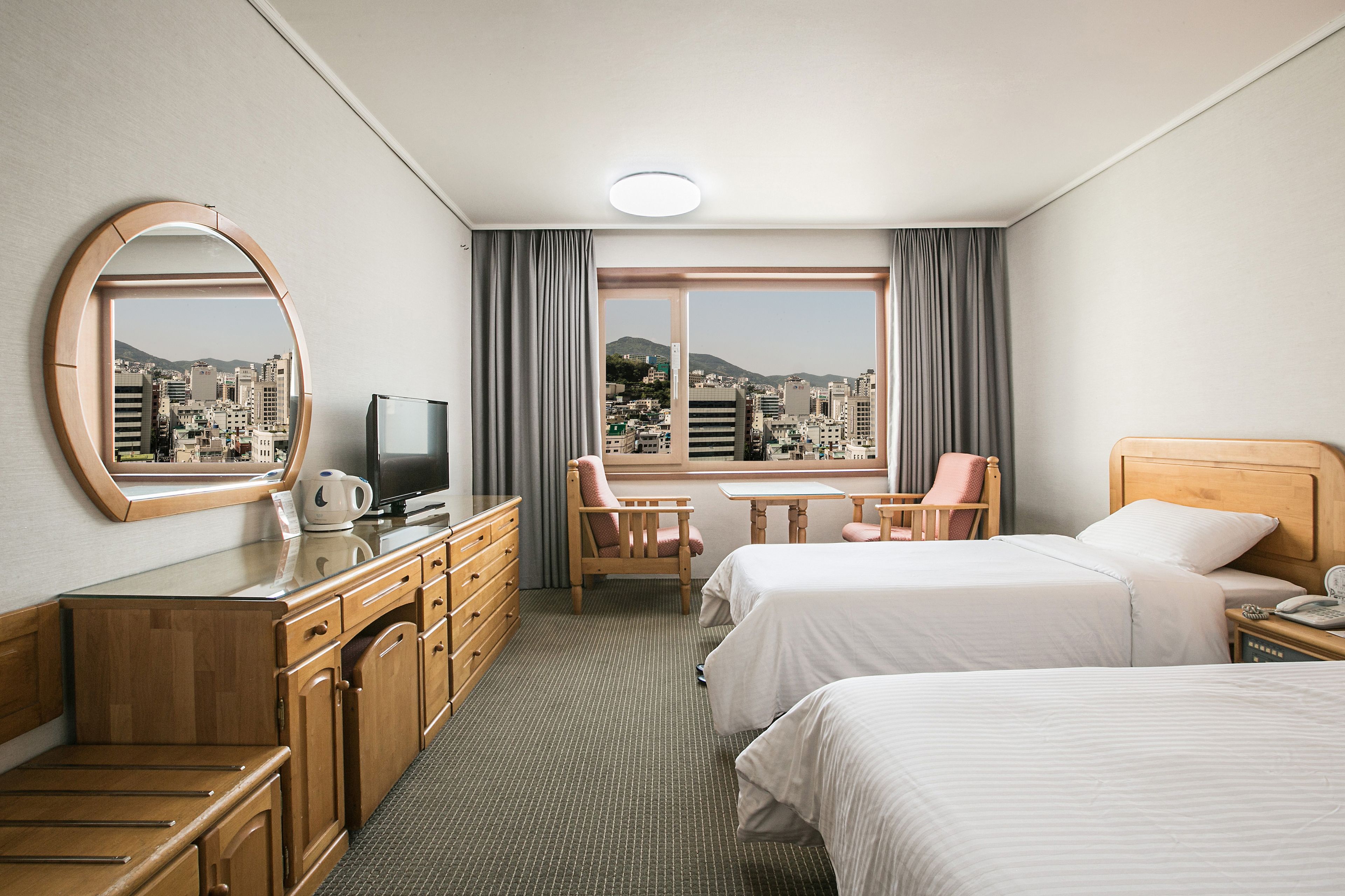 A hotel room with two beds, a TV, a desk with a chair, and a view of a cityscape through a large window.