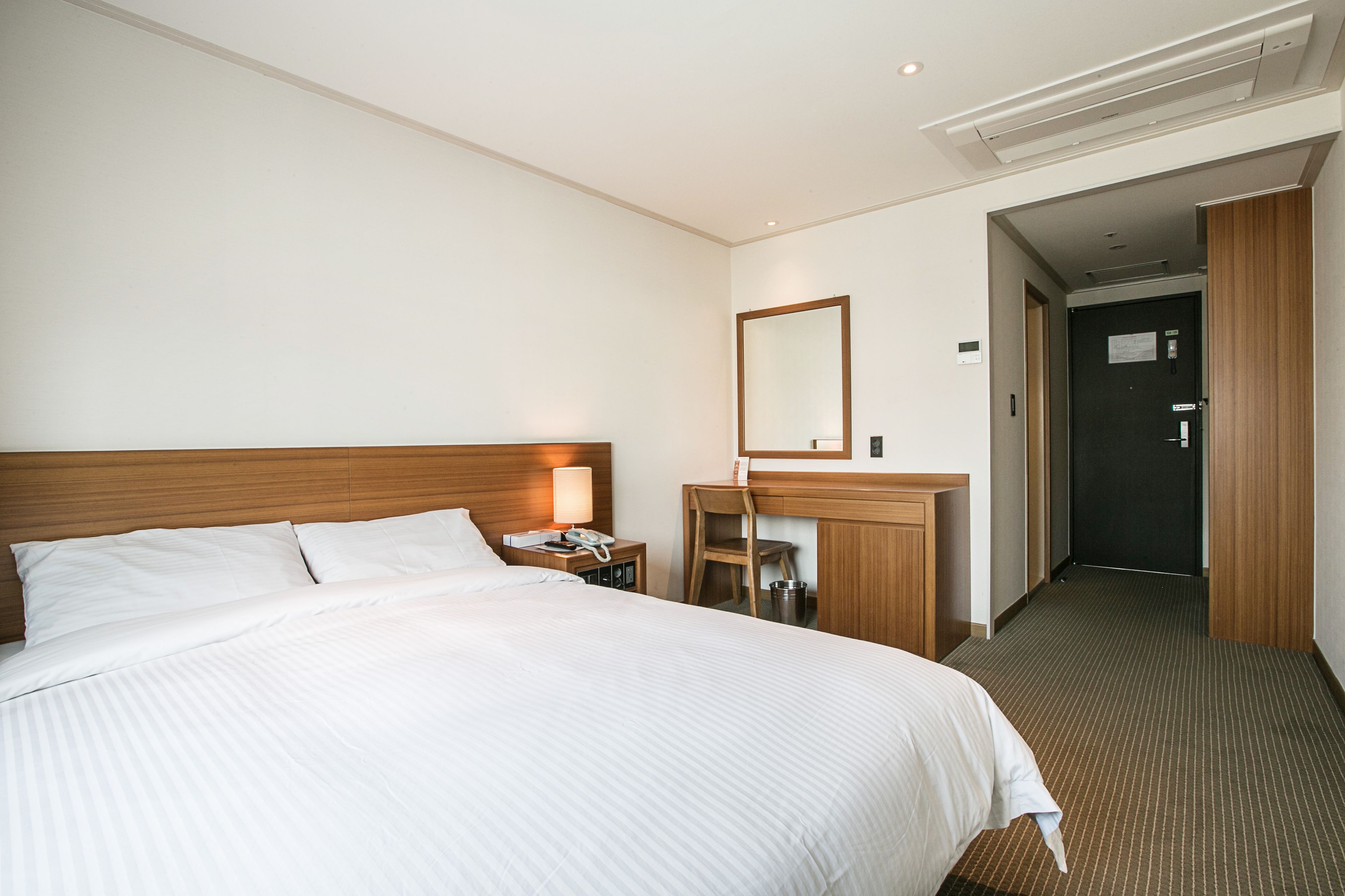 A hotel room with a large bed, a wooden headboard, a desk with a chair, a mirror, and a door leading to another room.