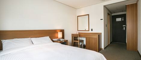 A hotel room with a large bed, a wooden headboard, a desk with a chair, a mirror, and a door leading to another room.