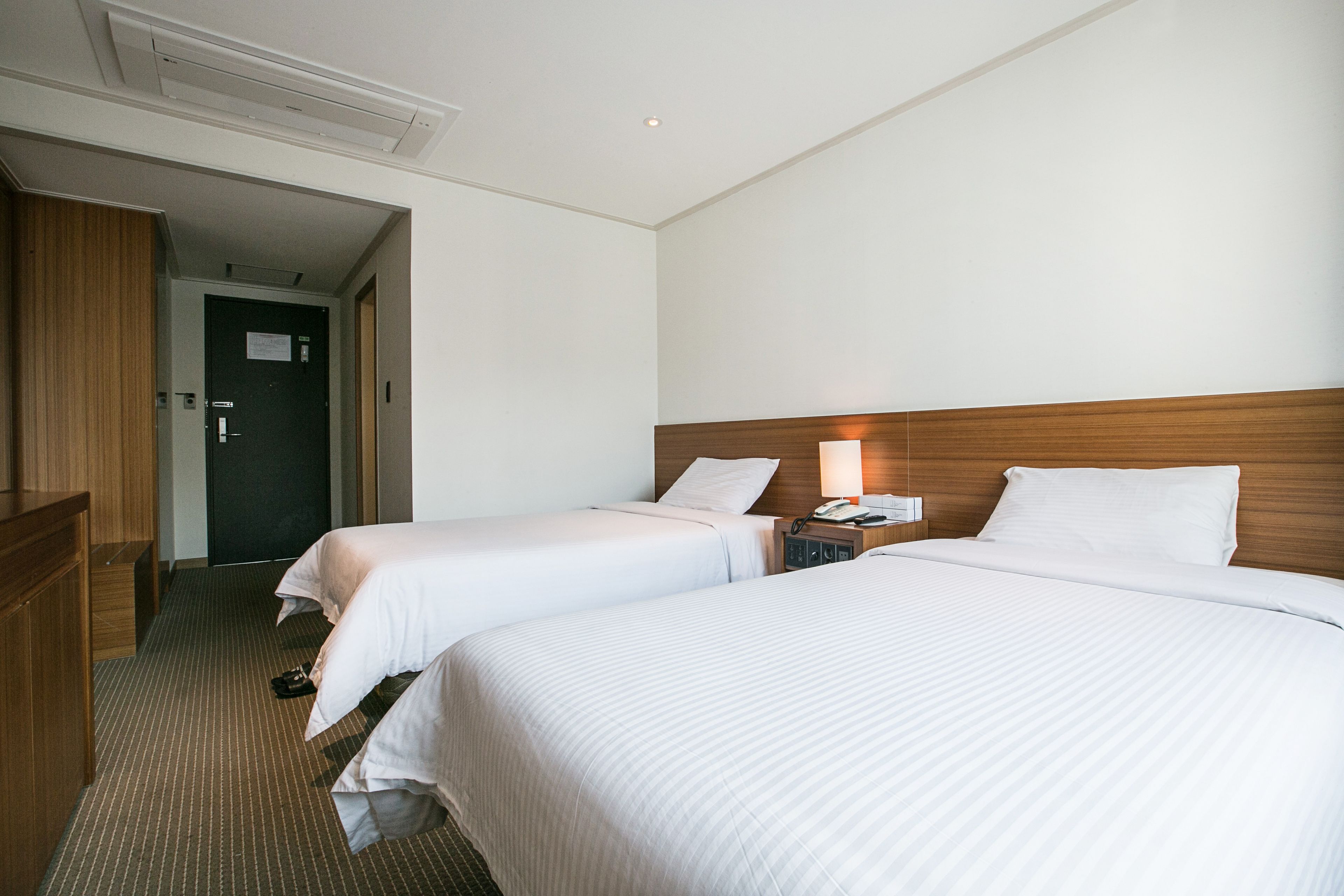 A hotel room with two beds, a wooden headboard, a nightstand with a lamp, and a door leading to another room.