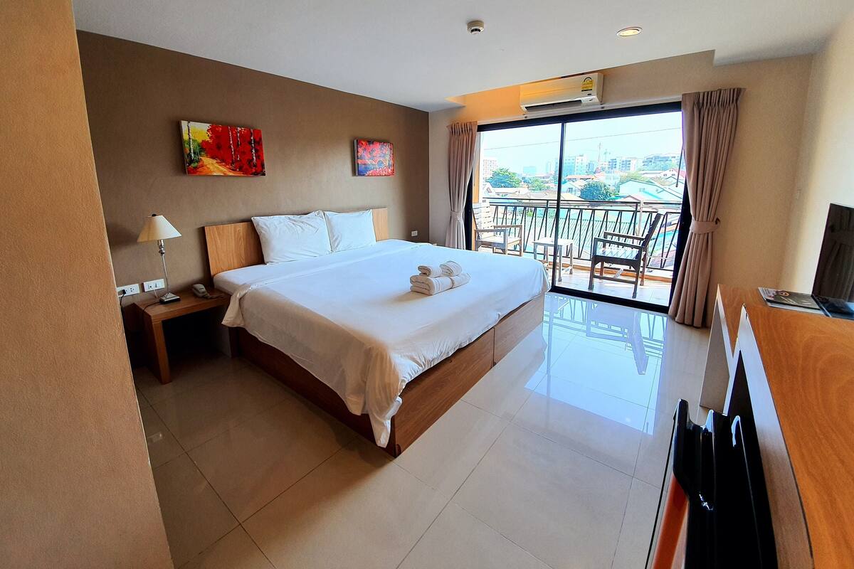 A hotel room with a large bed, a desk with a computer, a TV, and a balcony with a view of the city.