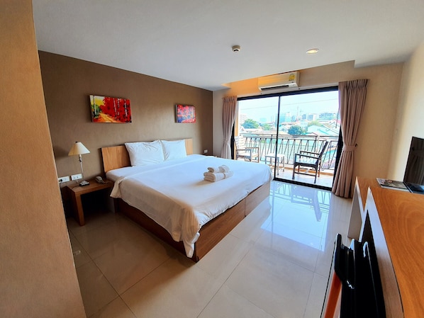 A hotel room with a large bed, a desk with a computer, a TV, and a balcony with a view of the city.
