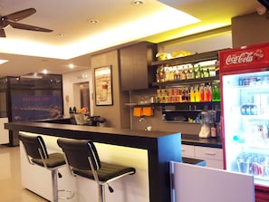 A bar area with a counter, two bar stools, a refrigerator stocked with beverages, and shelves with bottles.