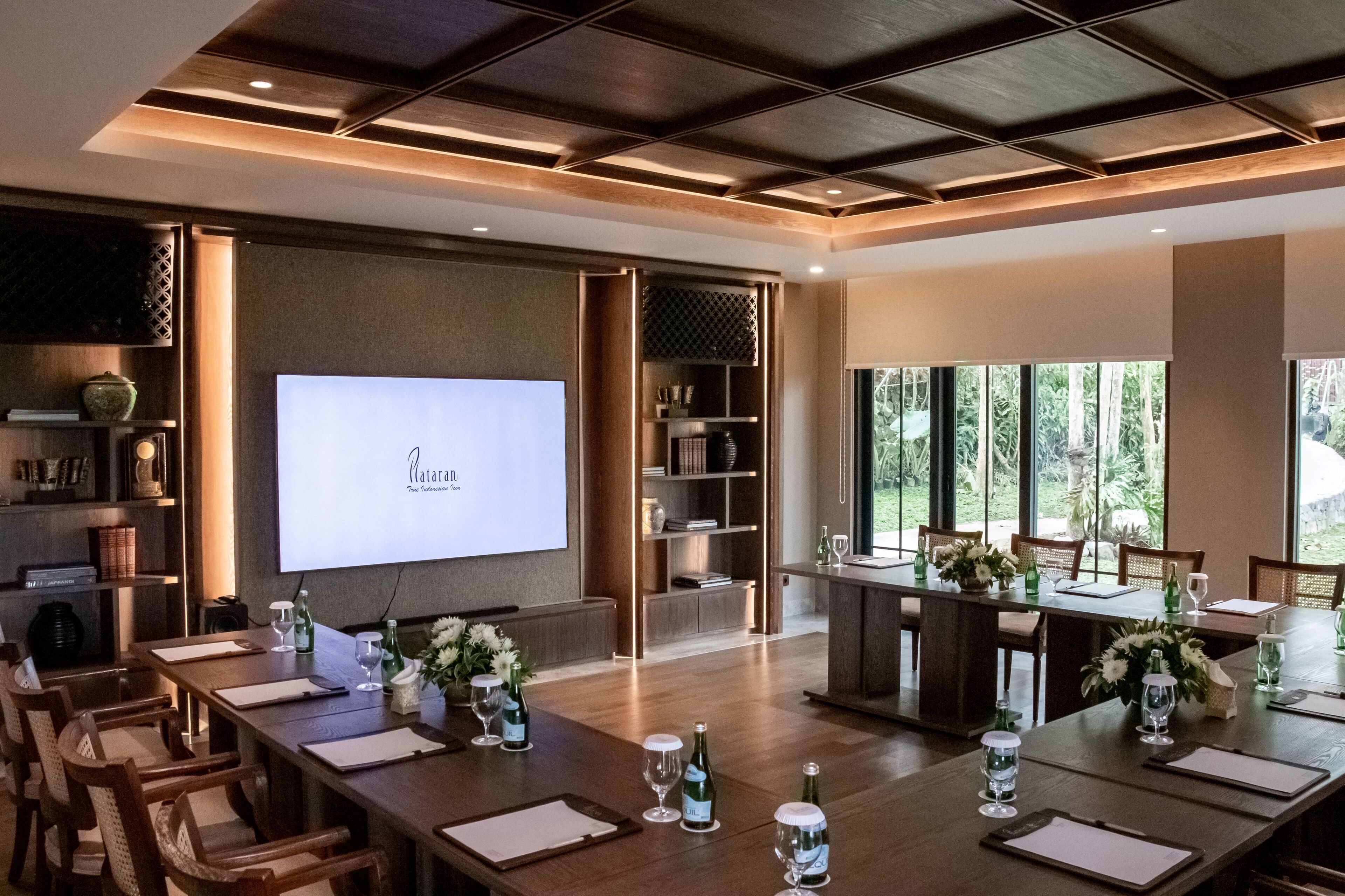 Sala de conferência com tela grande exibindo texto, mesas de madeira com cadeiras e vista para o verde lá fora.
