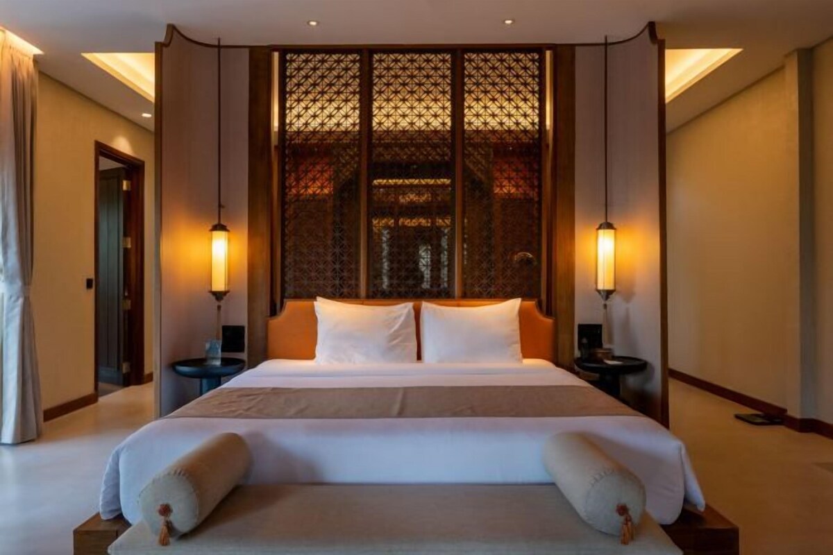 Habitación de hotel con una cama grande, dos mesitas de noche, un separador decorativo y dos lámparas de pared.