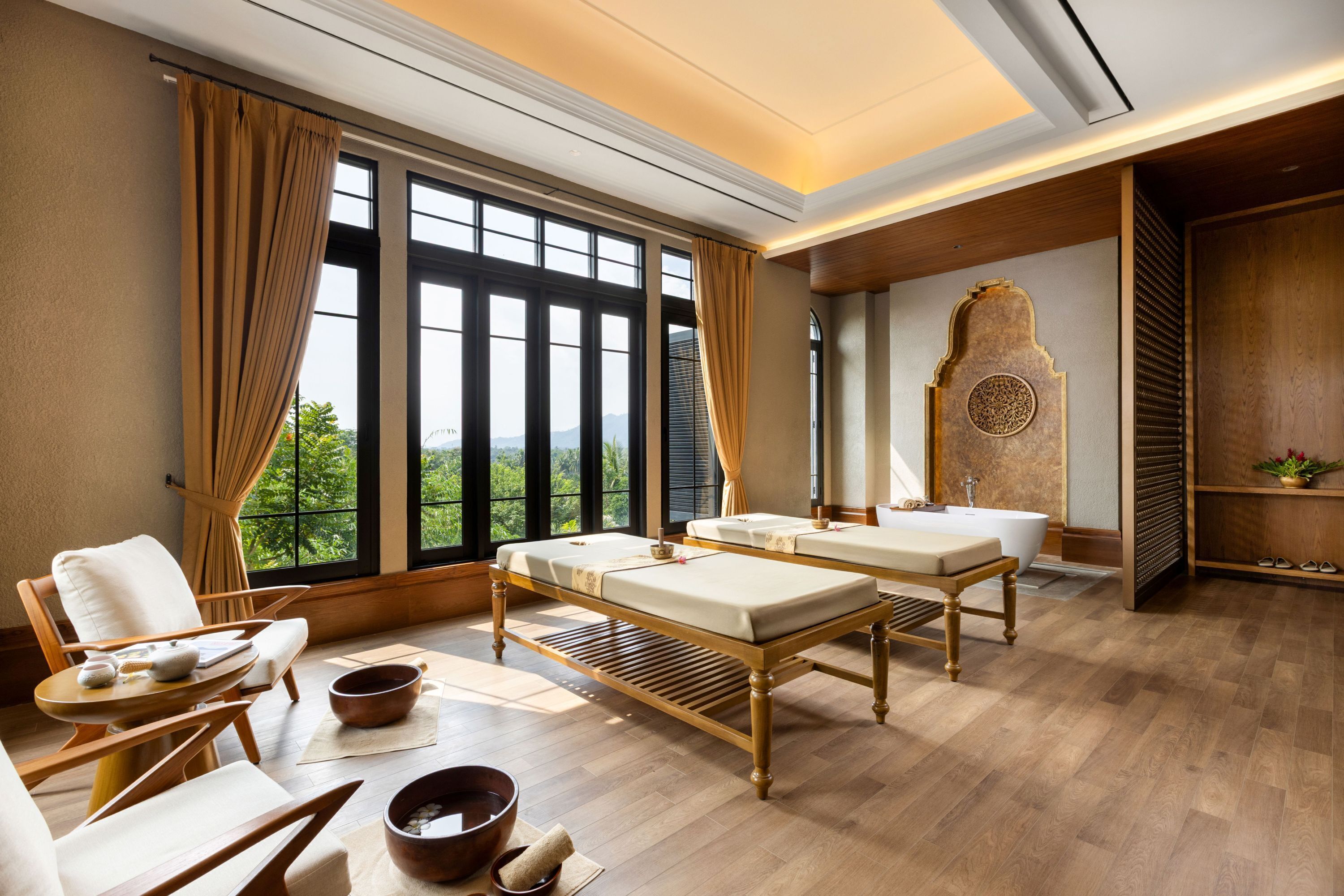 A spa room with a large window, a massage table, a chair, and a wooden cabinet.