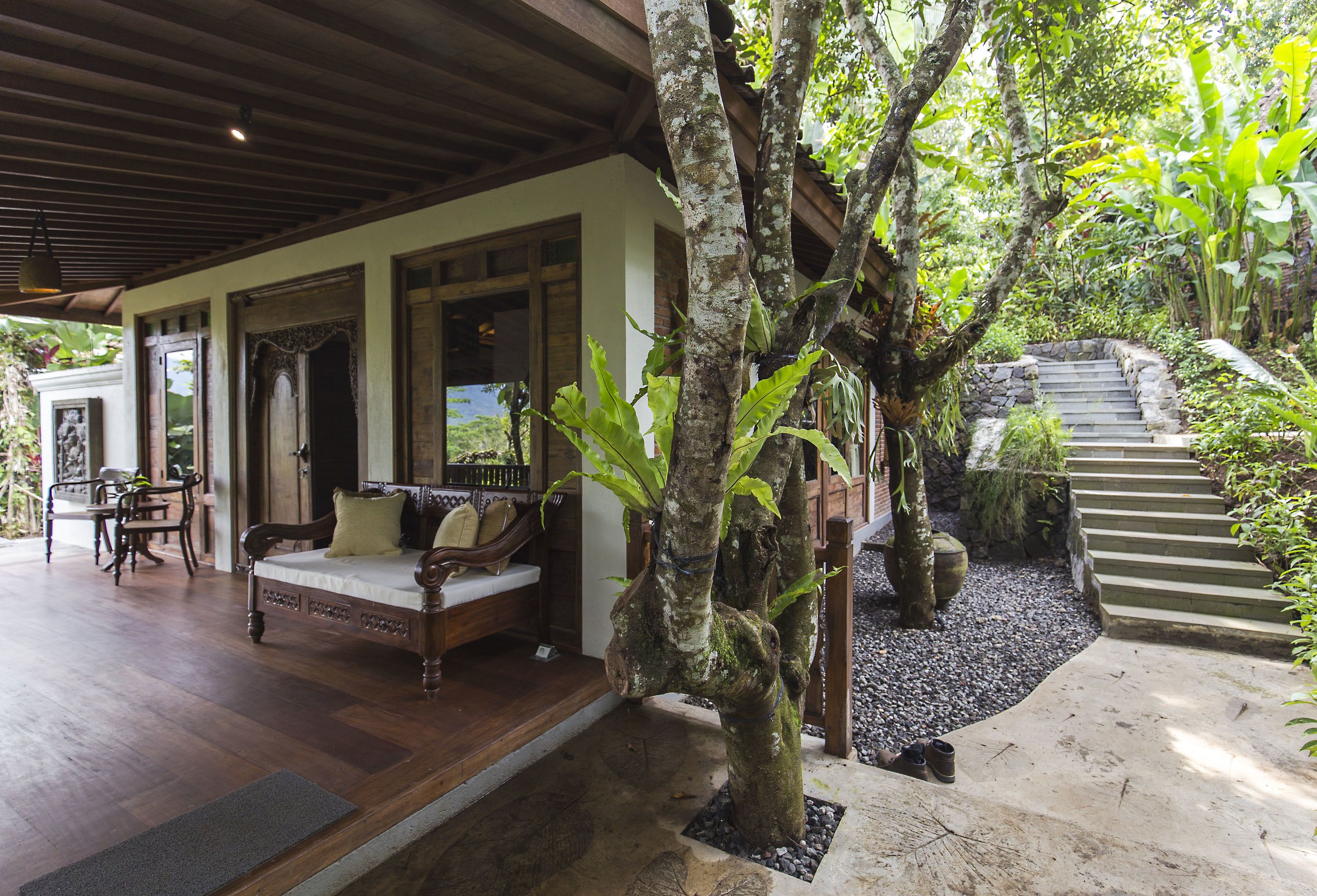 A covered outdoor seating area with a wooden bench and cushions, surrounded by lush greenery and a stone staircase leading to a higher level.