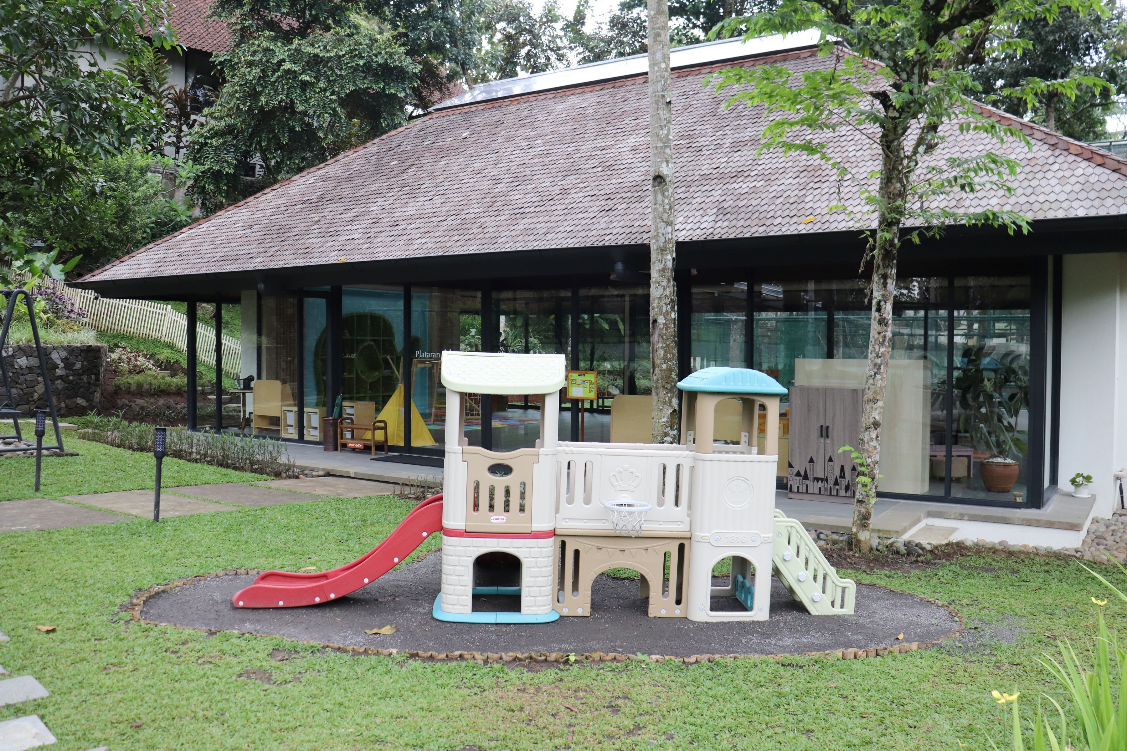 A children's play area with a slide and climbing structures, surrounded by a well-maintained lawn and a building with large windows in the background.