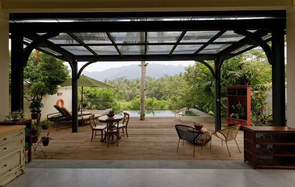 A covered patio with wooden flooring, outdoor furniture, and a view of lush greenery and mountains.