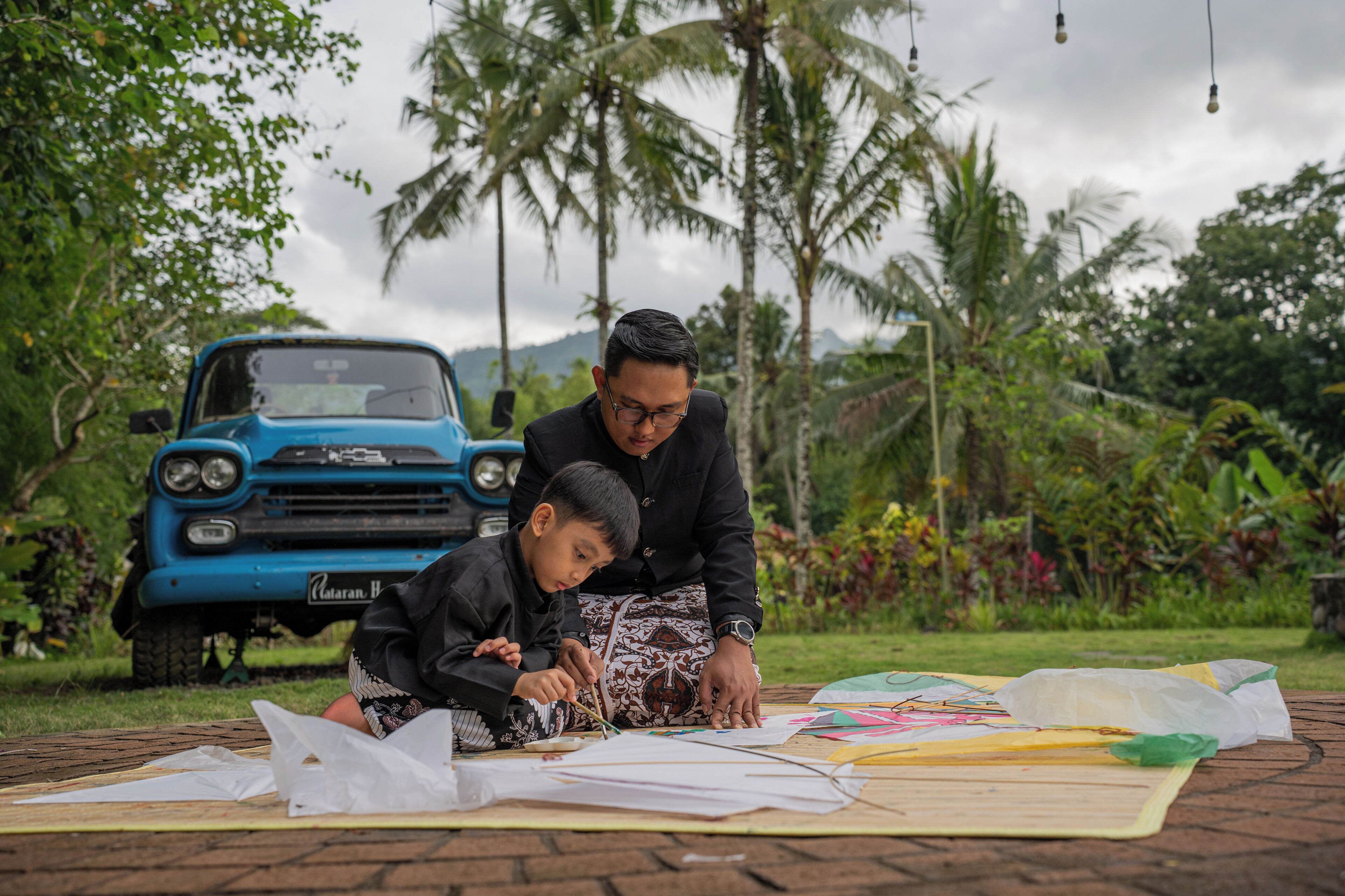 A man and a child sitting on a table outdoors with a blue vehicle in the background.