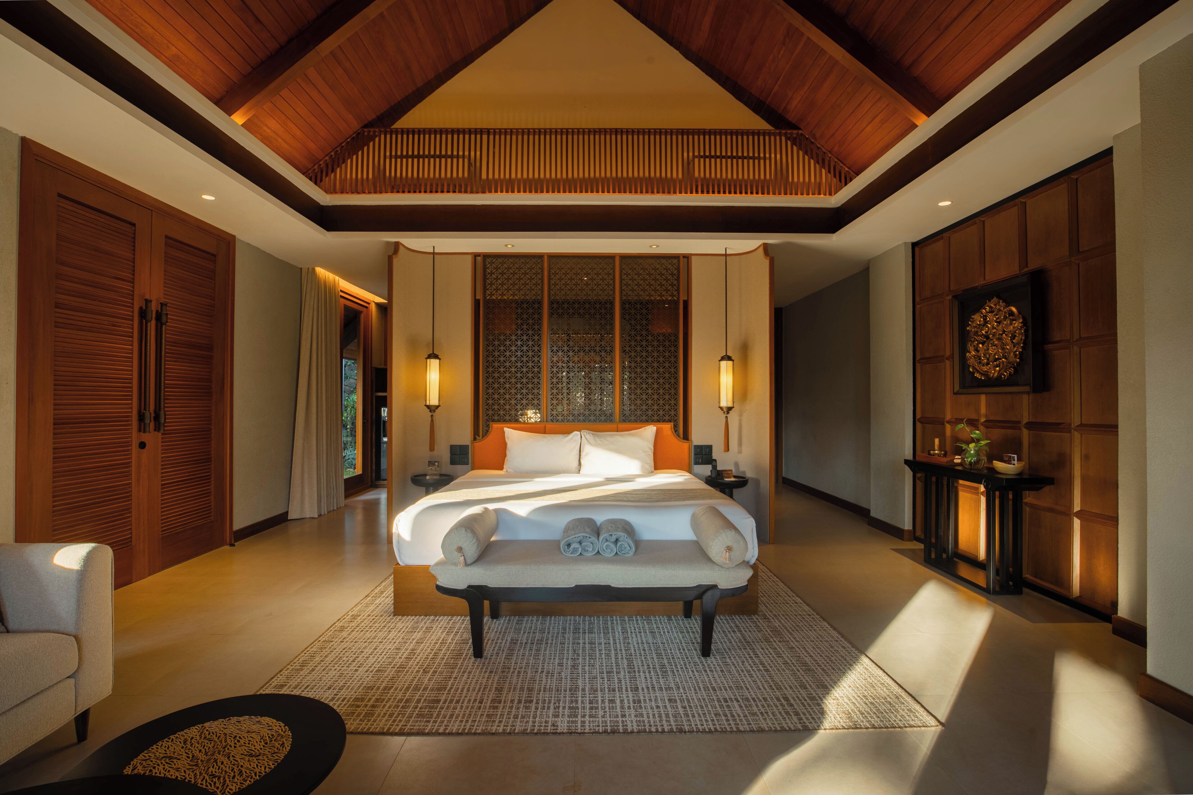 A spacious bedroom with a large bed, wooden ceiling beams, and a view of the outdoors through a sliding door.