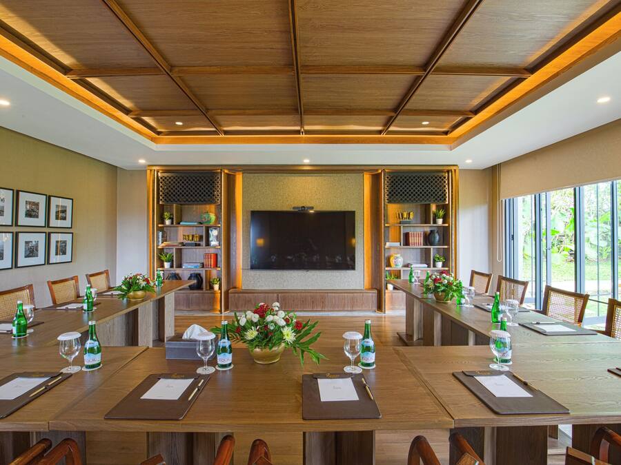 A conference room with a long table, chairs, a large screen, and shelves with books and decorative items.