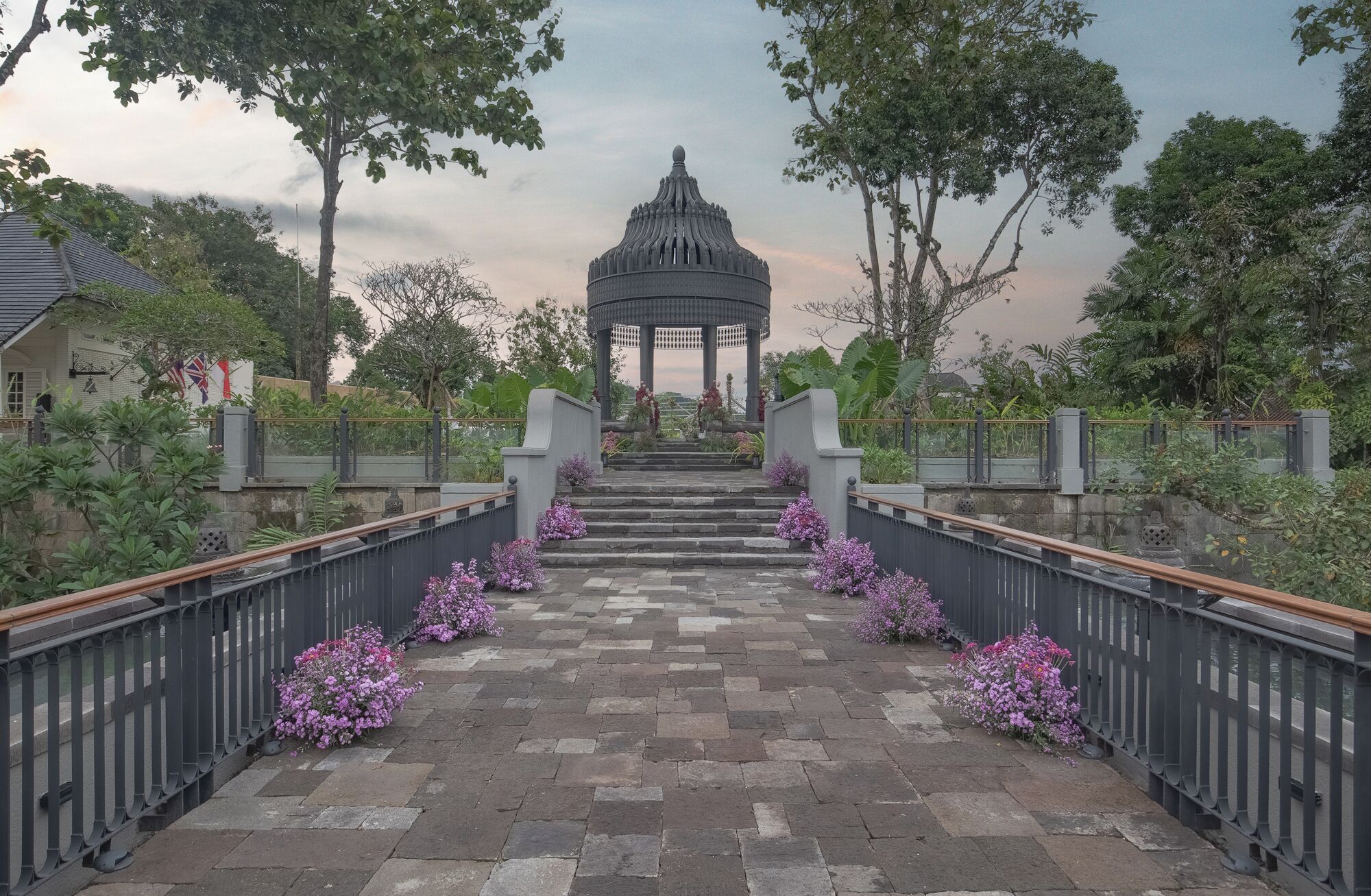 A stone pathway leading to a gazebo with a domed roof, surrounded by a metal railing and purple flowers.