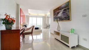 A modern hotel room with a wooden desk, a chair, a white console table, a painting of a waterfall, and a view of the outdoors through large windows.