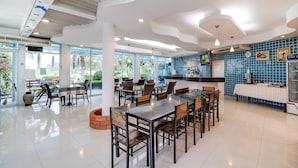 A spacious dining area with multiple tables and chairs, large windows offering views of the outdoors, and a kitchen area with blue tiles and modern appliances.