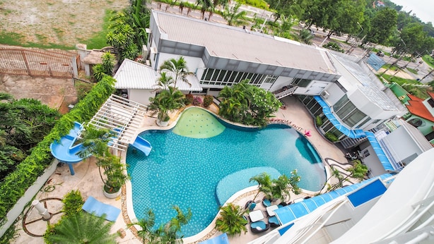 A swimming pool with a slide, surrounded by lounge chairs and palm trees, adjacent to a building with a sloped roof.