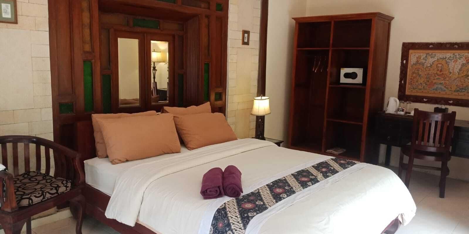 Una cama bien hecha con edredón blanco y dos almohadas marrones, una mesita de noche de madera con una lámpara, un armario de madera y una silla con cojín estampado.