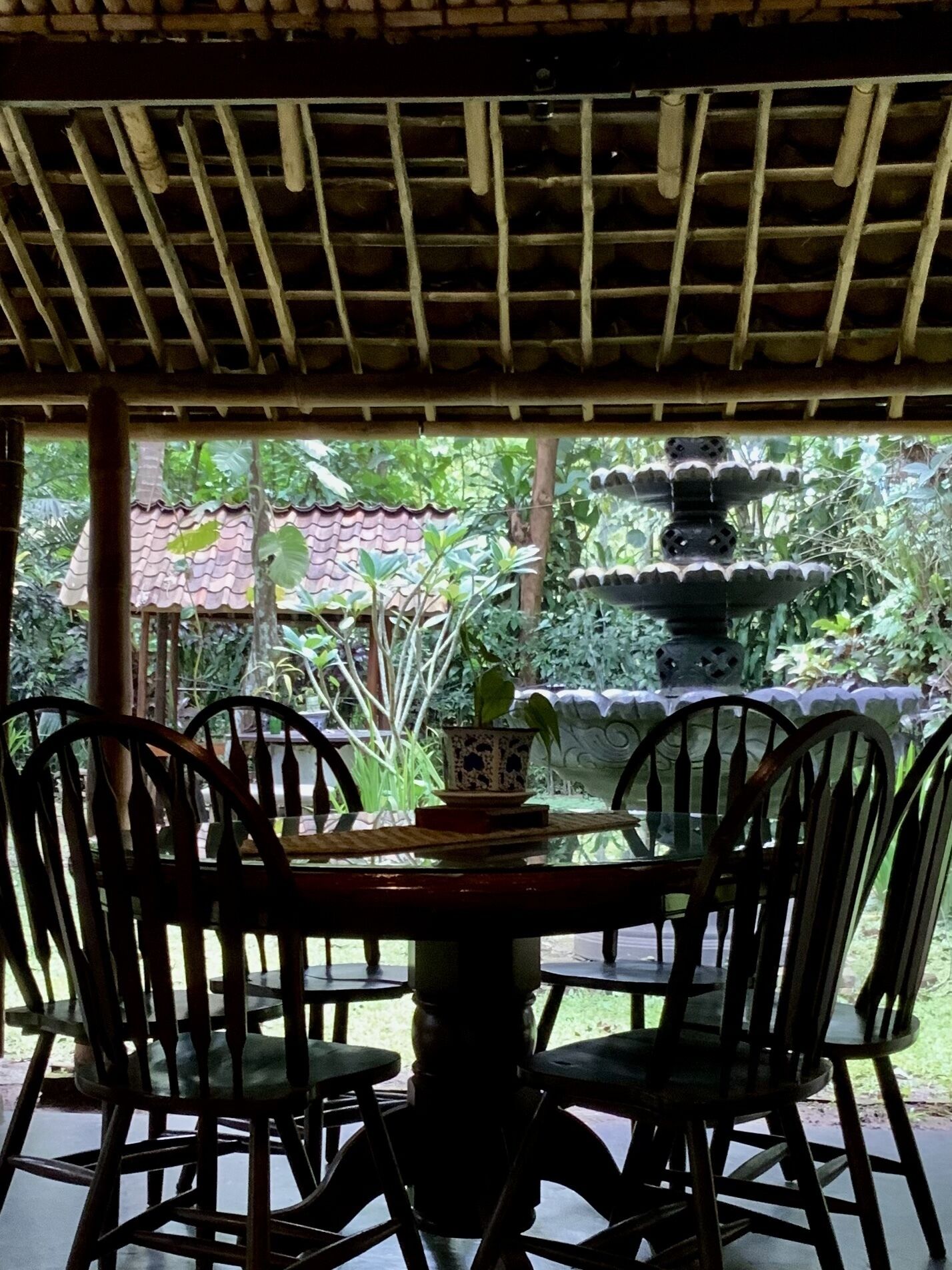 A covered outdoor dining area with a table and chairs, surrounded by lush greenery and a decorative fountain.