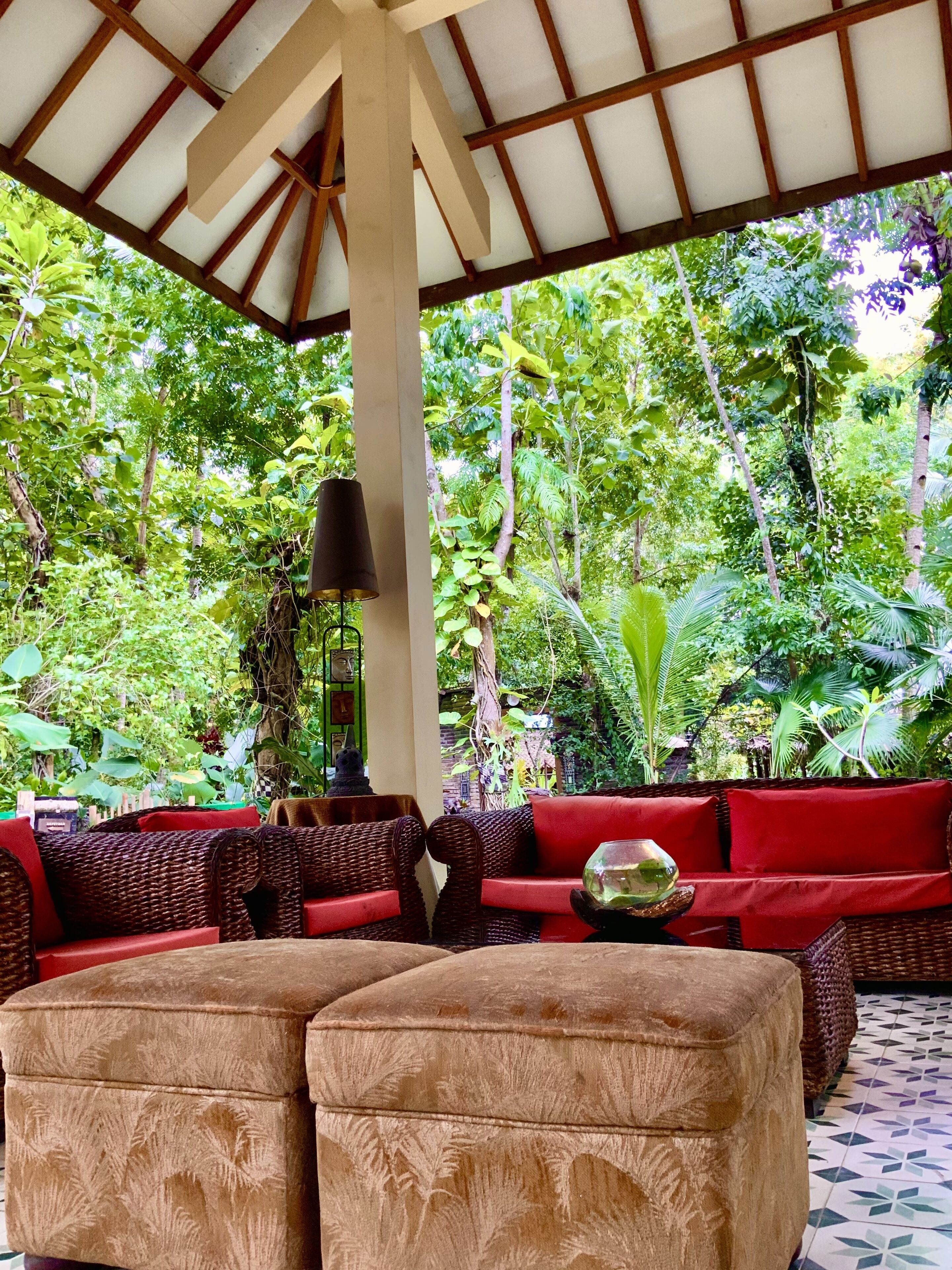 An outdoor seating area with wicker furniture and red cushions, surrounded by lush greenery.