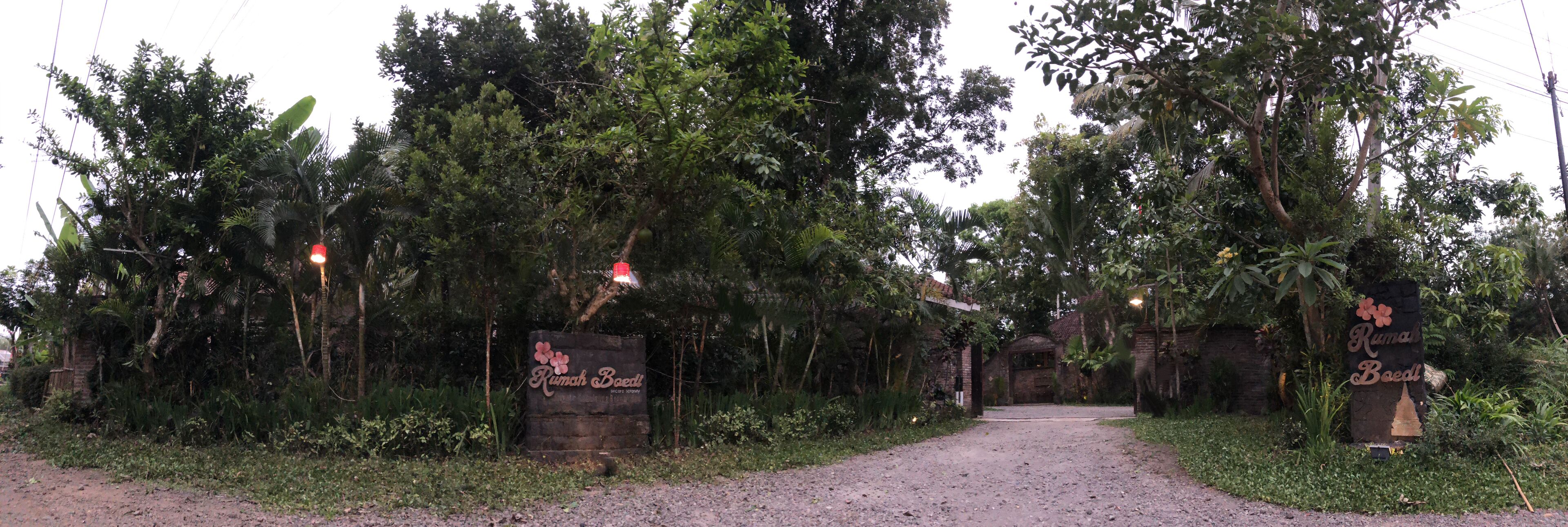 A rustic entrance with a gravel path leading through dense foliage, flanked by two stone signs with floral designs.