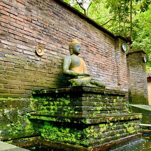 Eine Buddha-Statue auf einer moosbewachsenen Plattform vor einer Backsteinmauer.