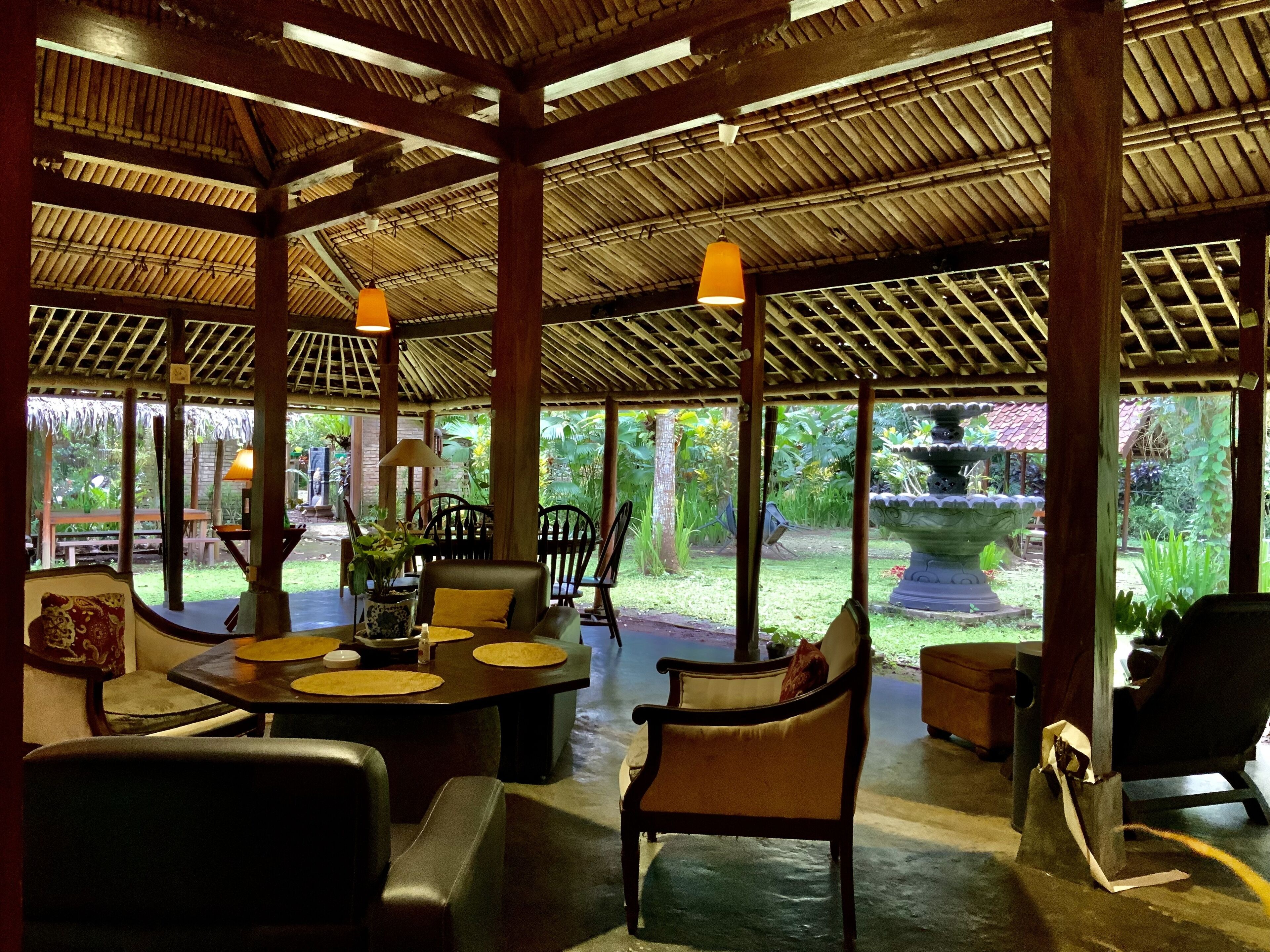 A covered outdoor seating area with tables and chairs, a thatched roof, and a view of a garden.