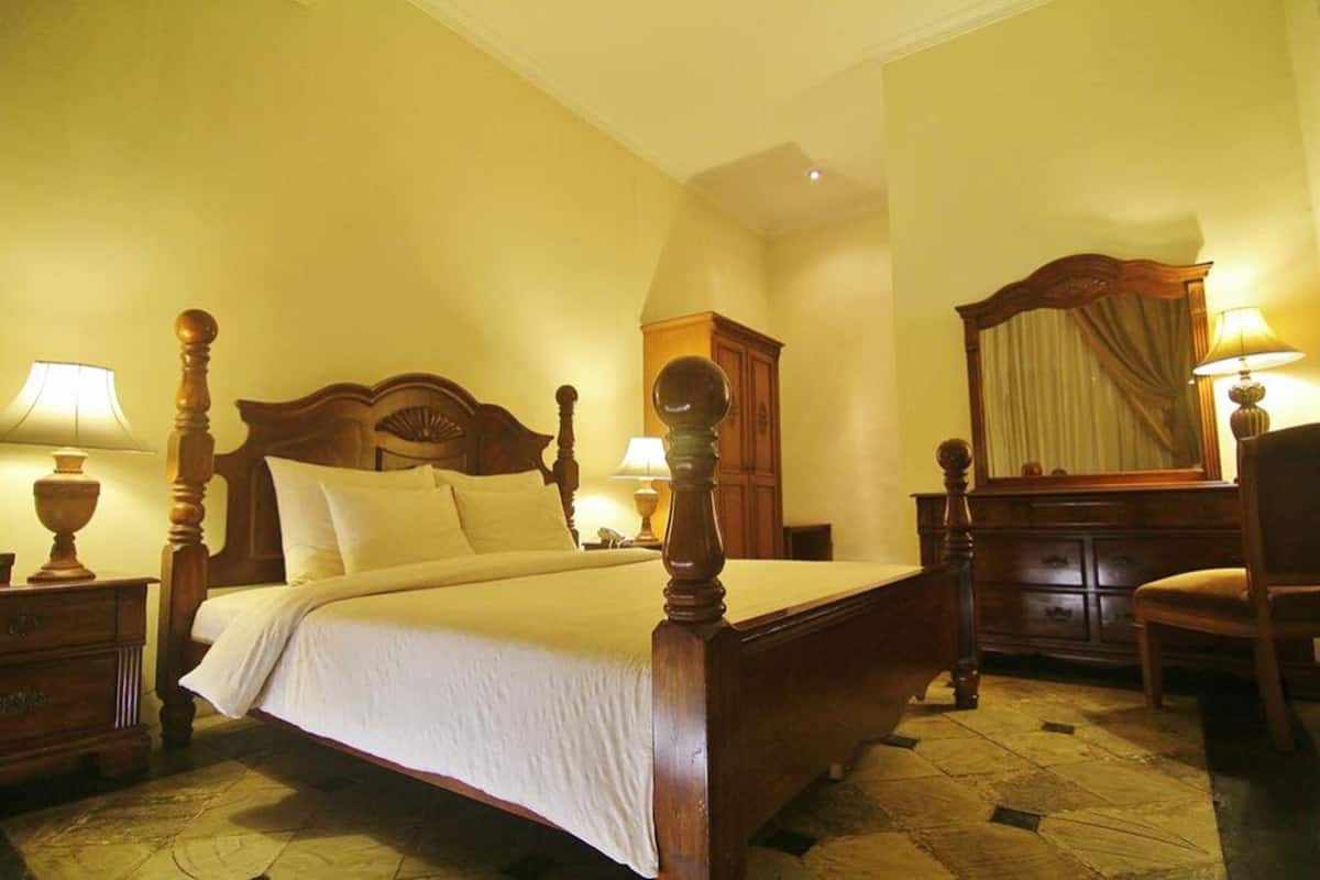 A traditional four-poster bed with a wooden headboard, flanked by two bedside tables with lamps. A wooden dresser with a mirror is to the right.