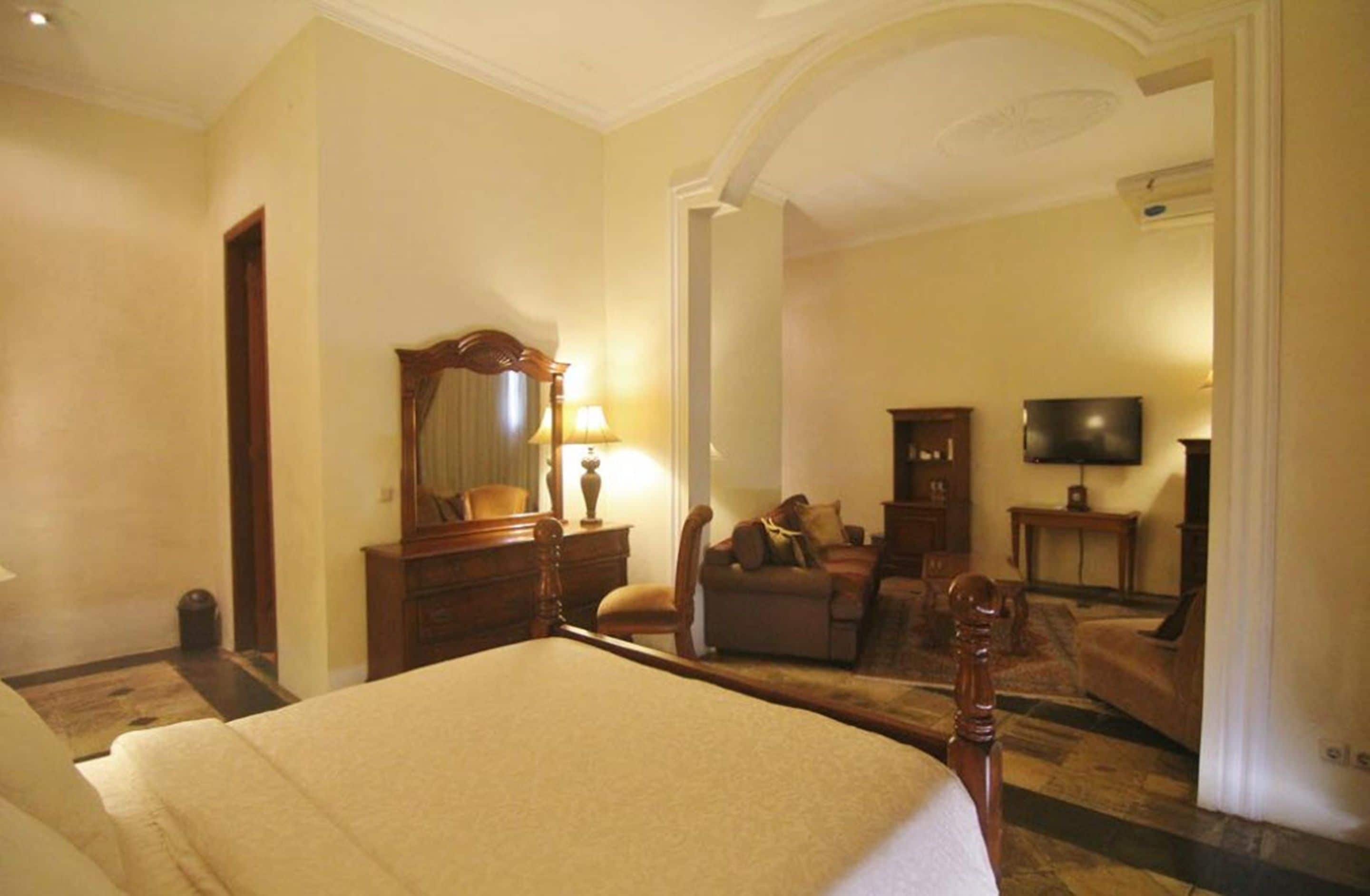 Habitación de hotel con una cama grande, un área de estar con sofá y sillones, un televisor y un espejo.