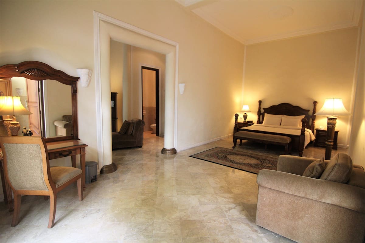 A spacious hotel room with a large bed, a sitting area with a sofa and armchair, a wooden desk, and a mirror.