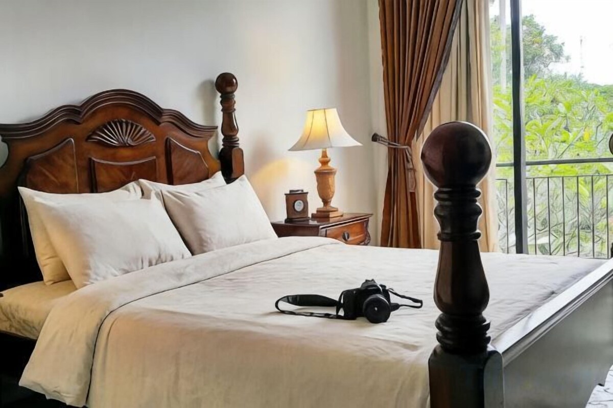 A neatly made bed with a wooden headboard, a bedside table with a lamp, and a view of greenery outside.
