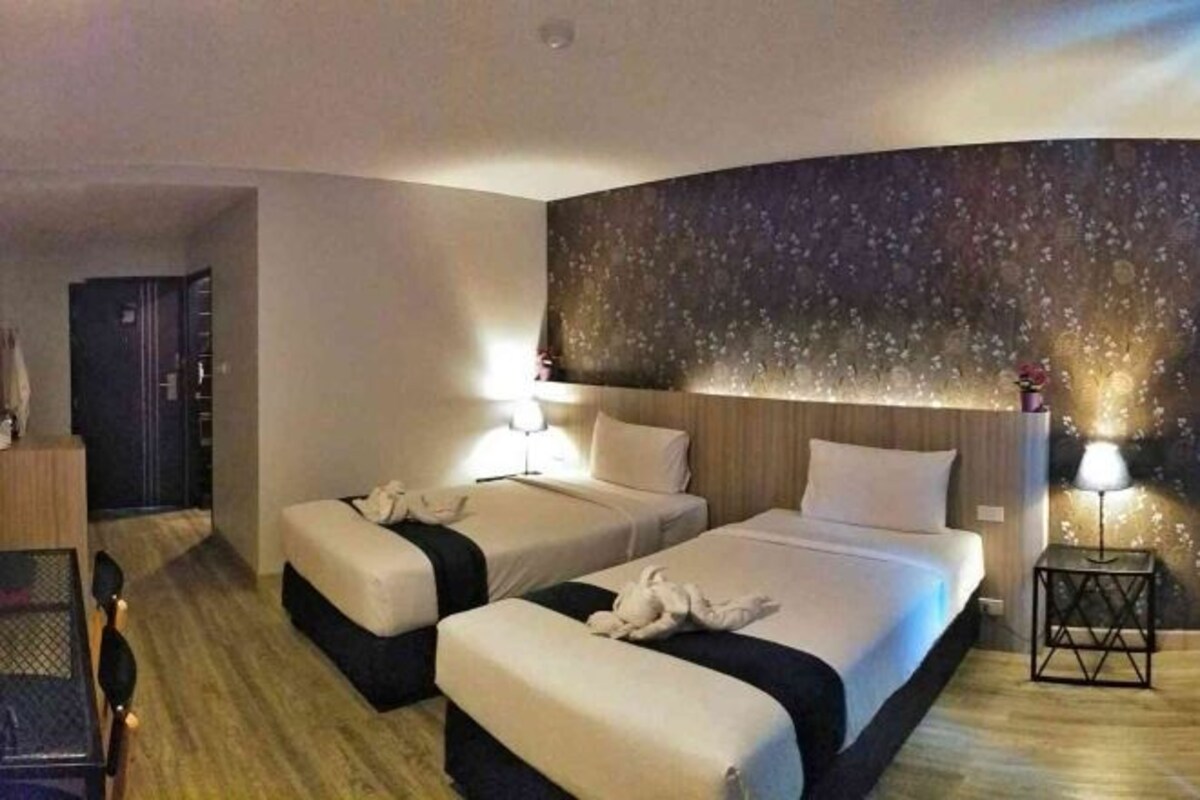 Una camera d'albergo con due letti, pavimento in legno, parete con motivo floreale e un tavolino con una lampada.