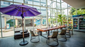 A spacious indoor area with a long wooden table and six wire-frame chairs, a purple umbrella, and a view of the outside through large windows.