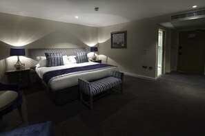 Une chambre d’hôtel avec un grand lit, deux tables de chevet, un banc et une porte donnant sur une autre pièce.