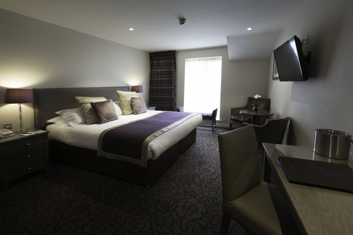 A hotel room with a large bed, bedside tables, a desk, a chair, a flat-screen TV, and a window with curtains.