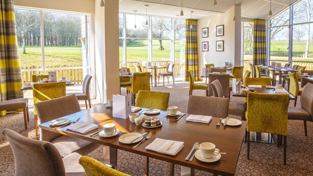 A dining area with tables set for four, chairs, and a view of a grassy field through large windows. - Sandford Springs Hotel & Golf Club (Tadley)