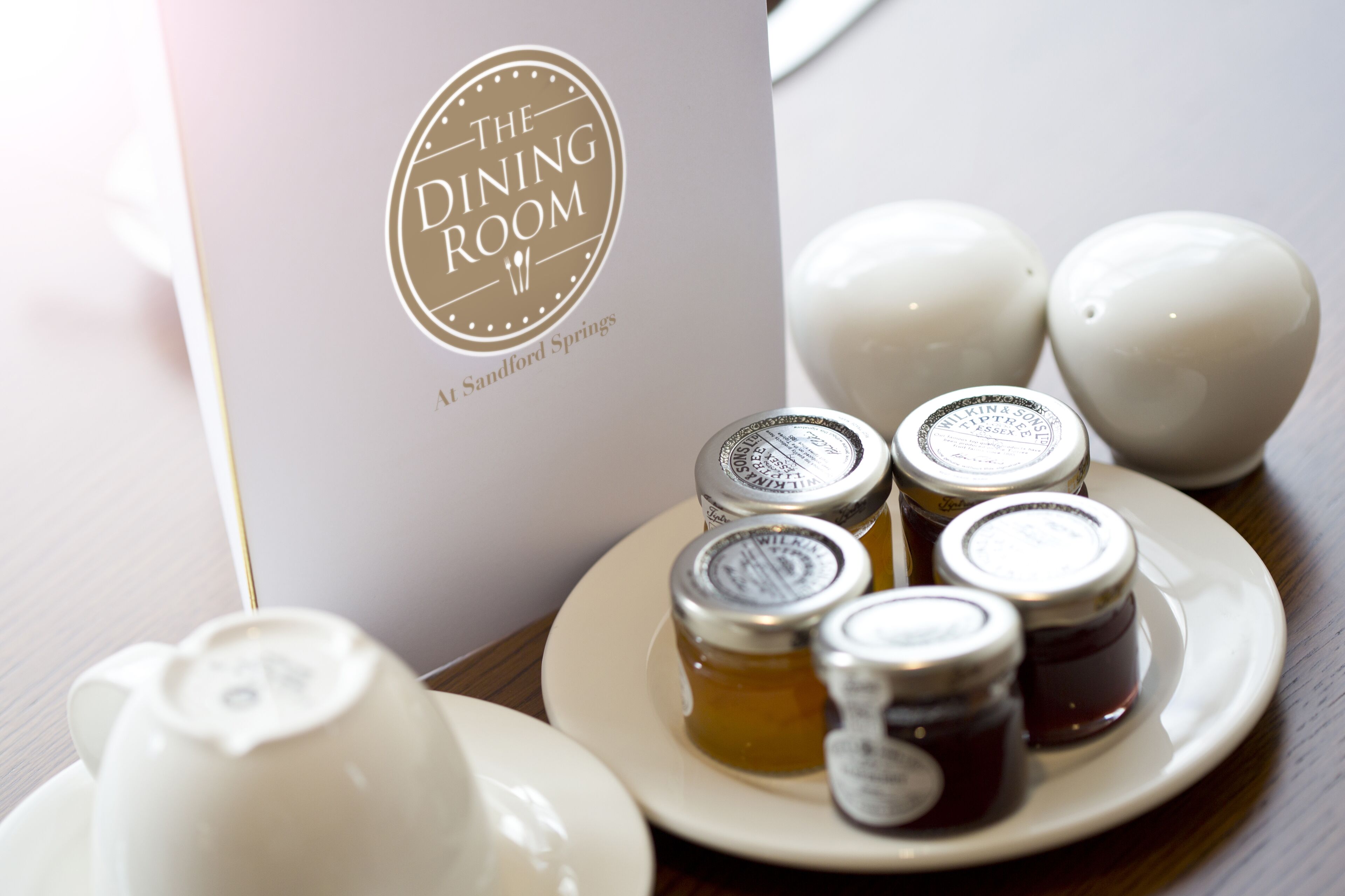 A table setting with a menu titled "The Dining Room" and four condiment jars on a plate.