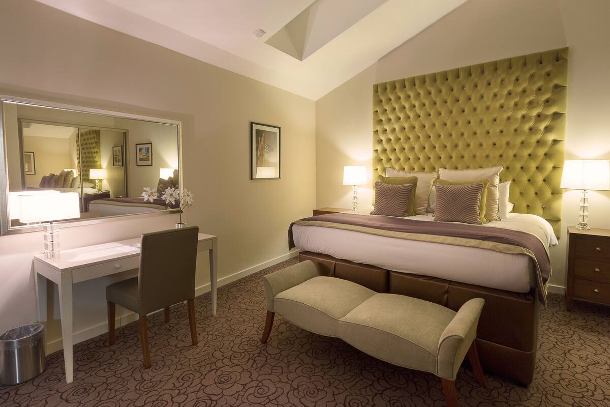 A hotel room with a large bed, a desk, a chair, and a bench.