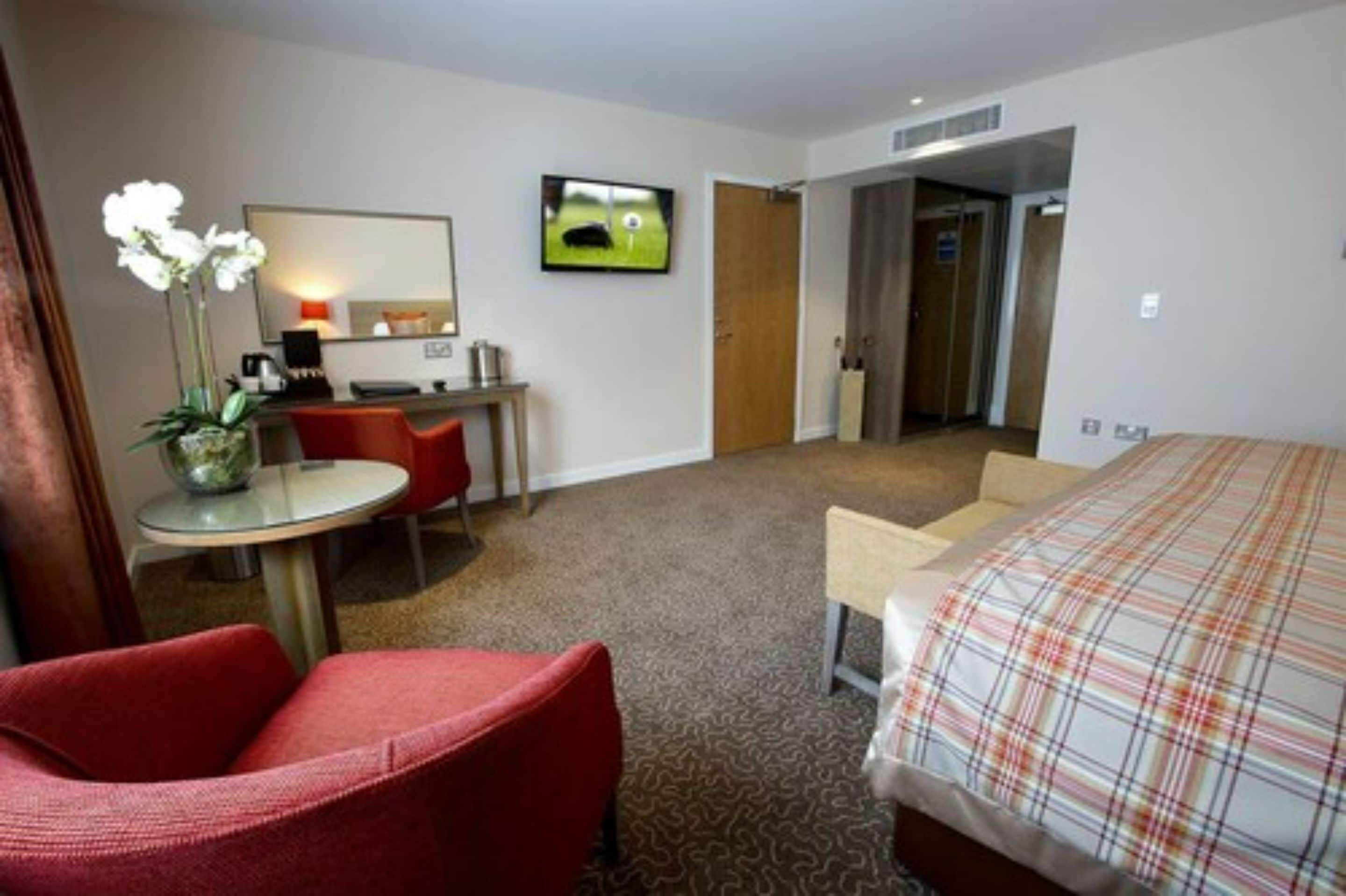 A hotel room with a bed, a desk with a chair, a TV, and a door leading to an elevator.