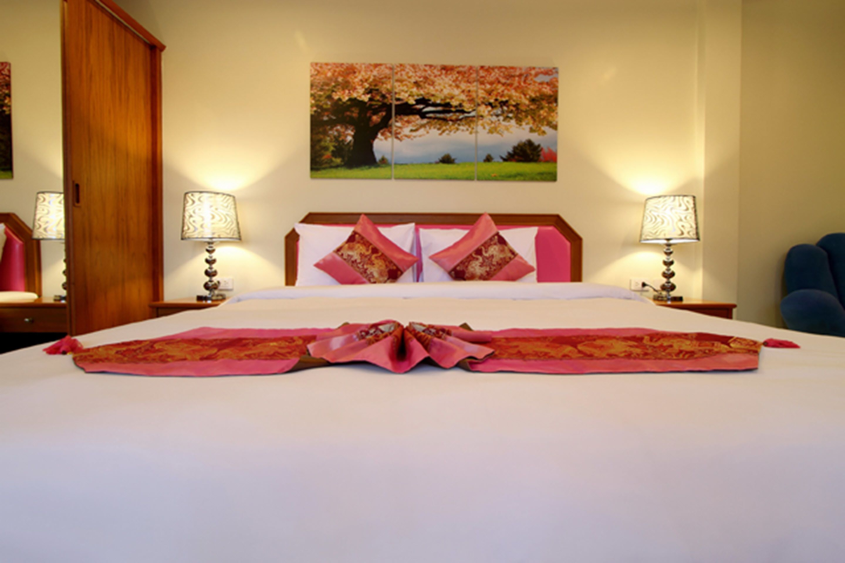 A hotel room with a large bed, two bedside lamps, a wooden headboard, a floral painting on the wall, and a blue chair.