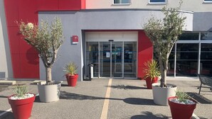 A building with a red and grey facade, a glass entrance, and potted plants in front. - Ace Hôtel Valence  (Valence)