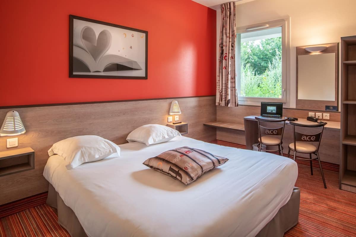 A hotel room with a bed, bedside lamps, a desk with a laptop, chairs, and a window with curtains.
