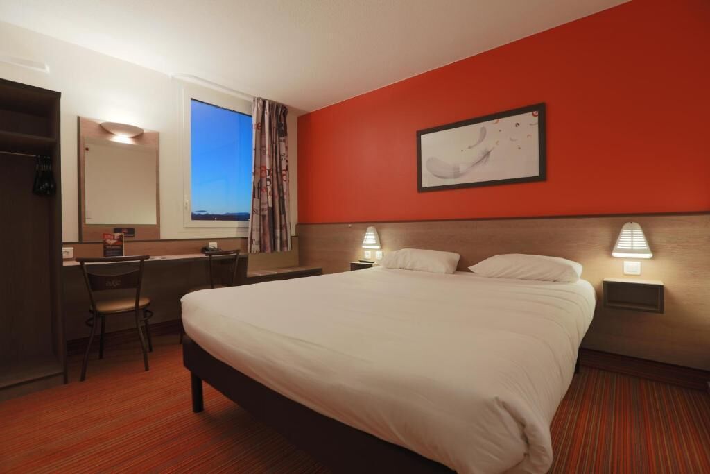 A hotel room with a large bed, a desk with a chair, a window with curtains, and a painting on the wall.