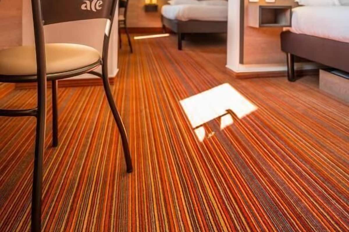 A hotel room with a striped carpet, a bed, a chair, and a nightstand with a lamp.