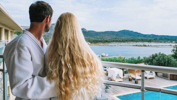 A couple on a balcony overlooking a pool with a view of a lake and mountains.