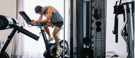 A person using a stationary bike in a room with a window, a wall-mounted rack, and a small table with a towel.
