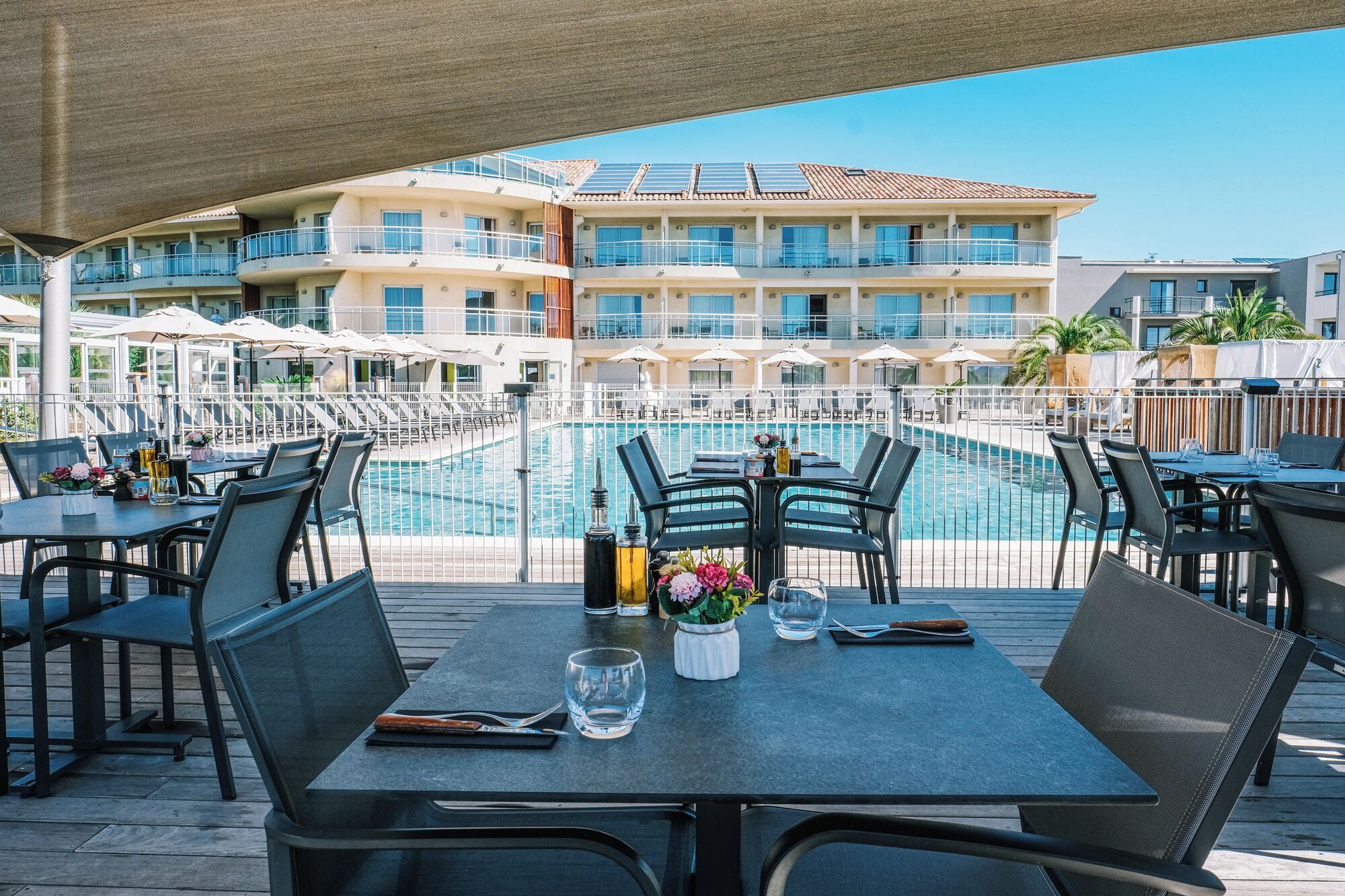 A poolside dining area with tables and chairs, a pool, and a building with multiple balconies in the background.