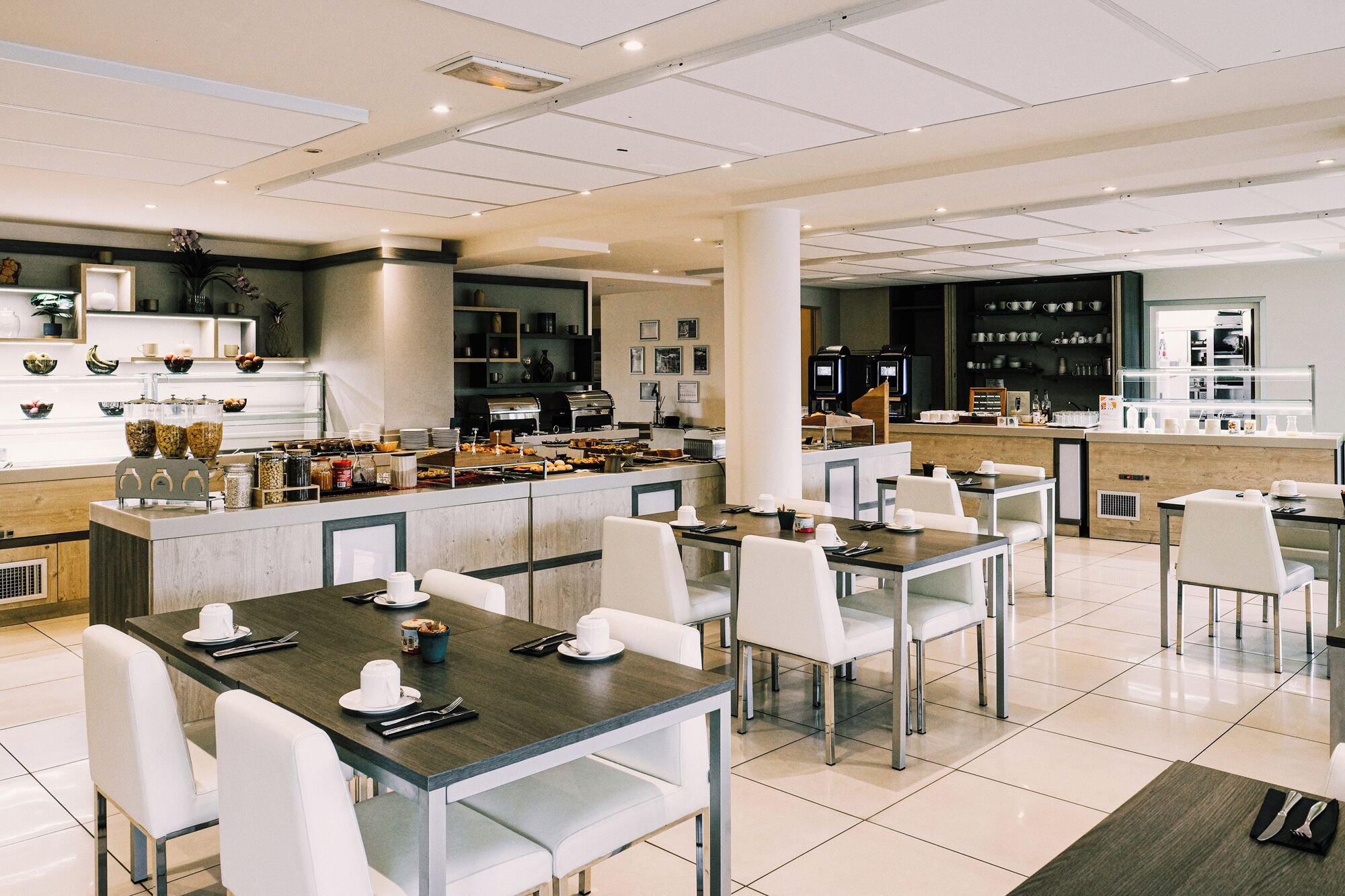 Een modern café met meerdere tafels en stoelen, een koffiebar en een ruim aanbod aan eten en drinken.