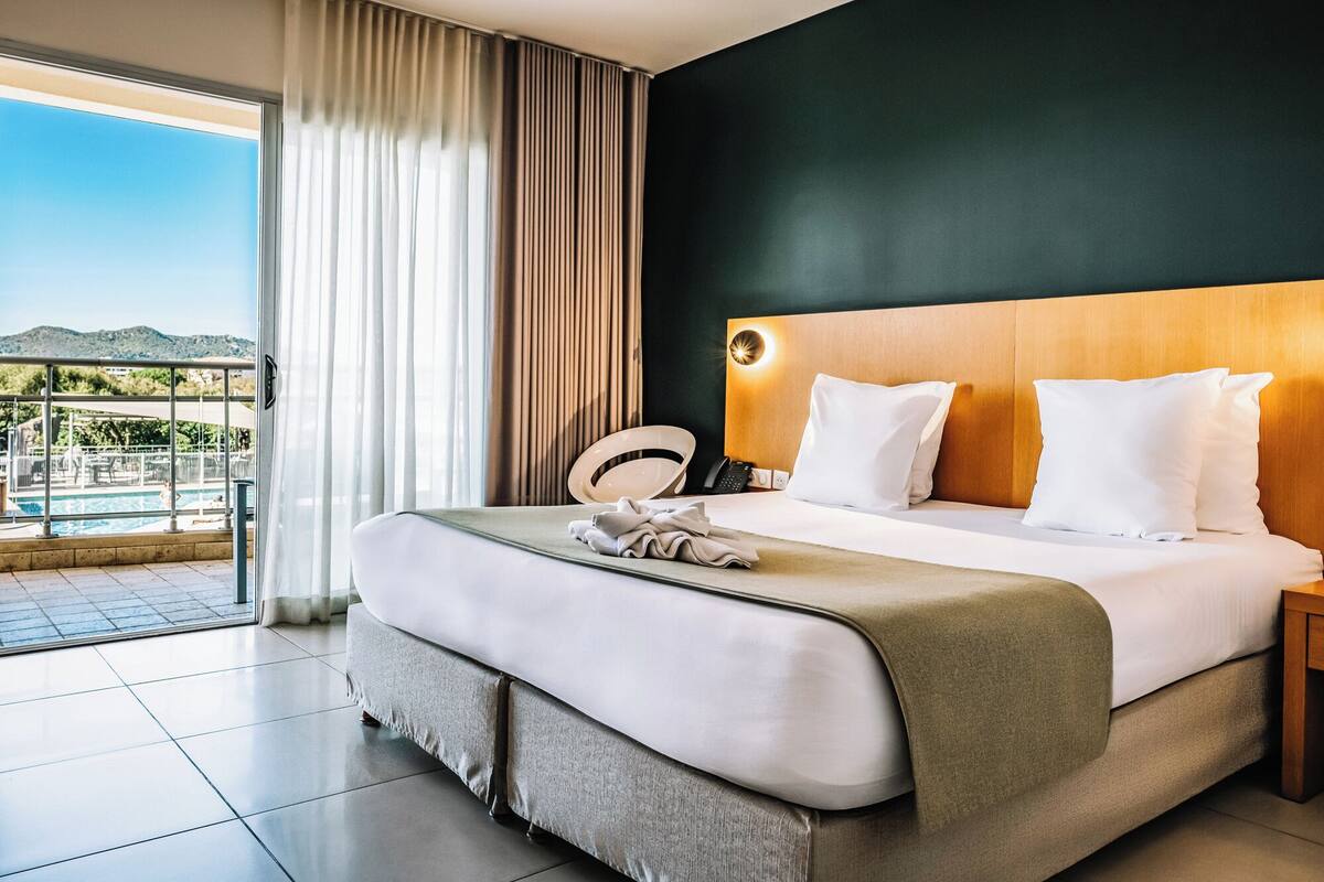 Une chambre d’hôtel avec un grand lit, une tête de lit en bois, une table de chevet avec une lampe, et une vue sur une piscine et des montagnes.