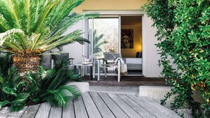 A modern outdoor seating area with a wooden deck, a large palm tree, and a glass door leading to a room with a bed and a painting.