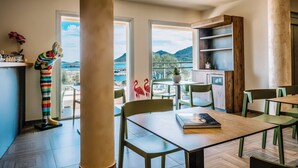 A modern dining area with a view of the sea and mountains, featuring a wooden table, green chairs, and a colorful sculpture.